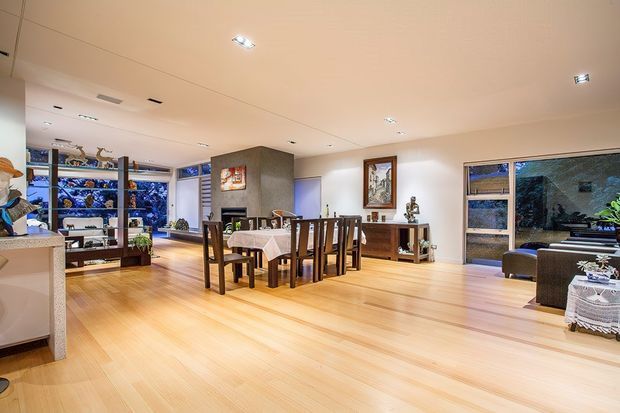 Open-plan interior with a dining area and adjacent living space. The room features warm light timber flooring, white walls and ceiling, recessed downlights, a dark timber dining table and chairs, a textured grey central fireplace or feature wall, built-in display shelving, and large windows overlooking greenery. Decorative objects and artwork add a traditional, homely character.