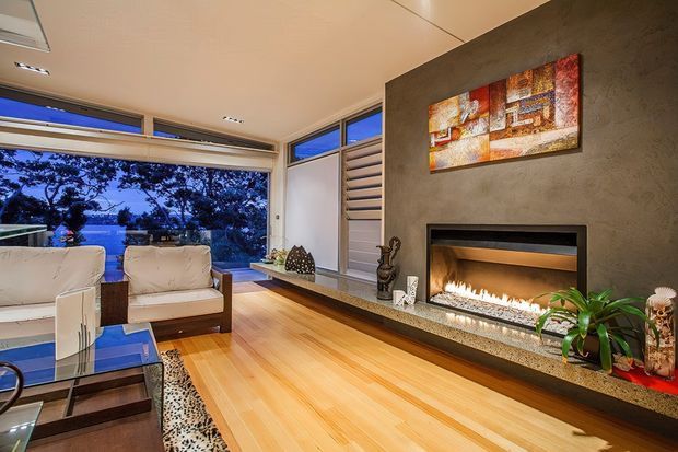 A refined contemporary living room with warm timber flooring, a large built-in linear fireplace set into a textured dark feature wall, and floor-to-ceiling glazing overlooking leafy surroundings. White lounge chairs, a glass coffee table, decorative artwork, and indoor plants create a comfortable, polished interior.