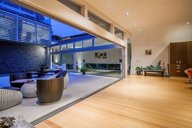 A spacious contemporary open-plan living area with pale timber flooring and a high sloping ceiling, opening through large glazed sliding doors to a covered outdoor courtyard. The room includes built-in dark timber cabinetry, a minimal console, indoor plants, and a glimpse of an outdoor lounge with a dark textured feature wall and horizontal screening. Recessed downlights and extensive glazing create a bright, indoor–outdoor connection.