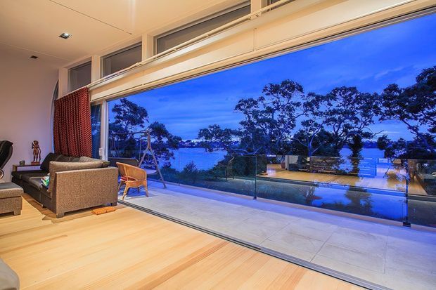 A contemporary living room with pale timber flooring, a neutral upholstered sofa, and full-height sliding glass doors opening onto a terrace with a glass balustrade. The view beyond shows mature trees, a swimming pool, and a calm waterfront setting at dusk. The room has a warm, open atmosphere with integrated ceiling lighting and red patterned curtains.