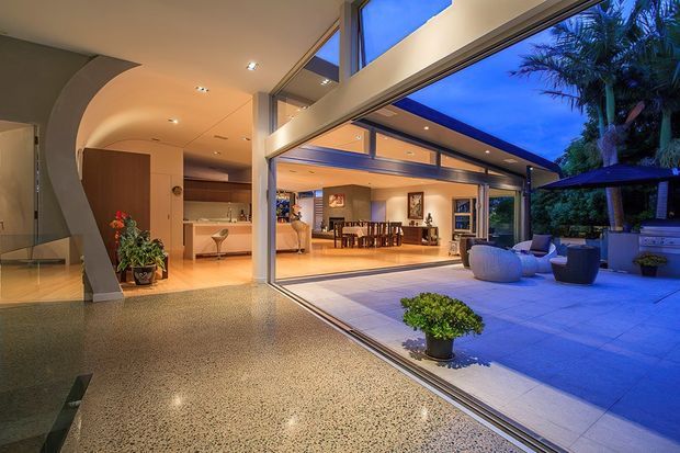 A sophisticated open-plan interior opening onto a covered outdoor terrace through expansive floor-to-ceiling sliding glass doors. The space features polished terrazzo flooring, warm recessed lighting, curved plastered architectural forms, timber cabinetry, and a dining area extending toward a landscaped courtyard and outdoor lounge.