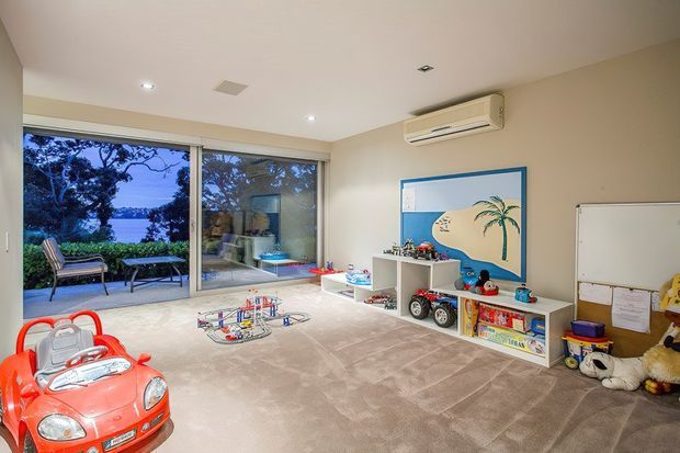Bright, spacious children's playroom with large sliding glass doors opening to an outdoor terrace, beige carpet, white walls, low open toy storage, a large wall-mounted pinboard, toys, and a red ride-on car. The room has recessed ceiling lights and a wall-mounted split-system air conditioner, with leafy outdoor views visible through the glazing.