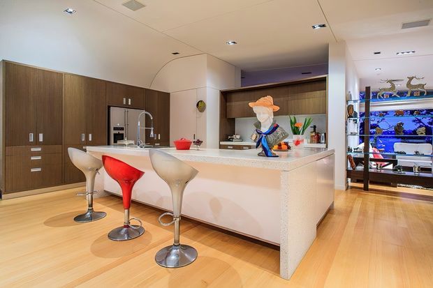 A bright contemporary kitchen with extensive dark timber cabinetry, a large pale stone island with waterfall sides, and four sculptural bar stools. The kitchen opens toward a display shelving area with illuminated aquarium-like blue lighting. Recessed ceiling lights and light timber flooring create a clean, modern interior.