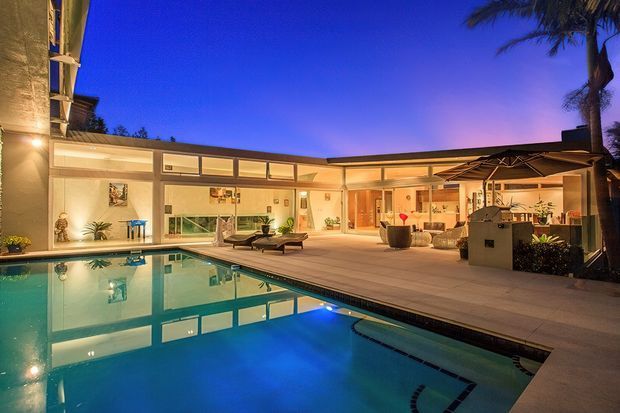 Twilight view of a modern single-storey home opening onto a large illuminated swimming pool and paved pool terrace. The building features extensive glazing, a low horizontal roofline, warm interior lighting, outdoor lounge furniture, and a covered outdoor entertaining area with a built-in barbecue or kitchen zone. Palm trees frame the scene.
