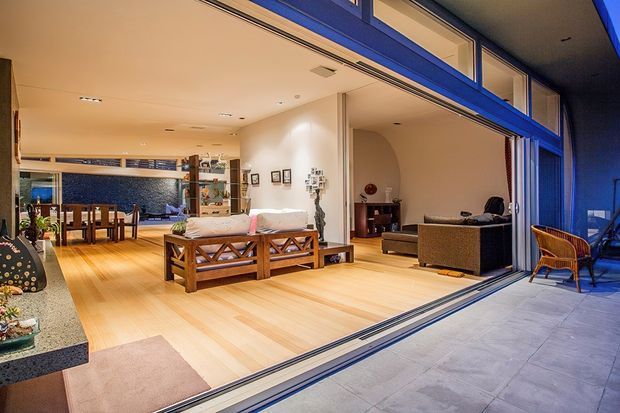 Open-plan residential interior viewed from a covered outdoor terrace through fully retracted blue-framed sliding glass doors. The bright living room has pale timber flooring, a white sofa, timber-framed coffee table, and a darker upholstered sofa, with a dining area and kitchen visible in the background. The space connects directly to the outdoor paved area and features warm recessed lighting and broad indoor–outdoor glazing.