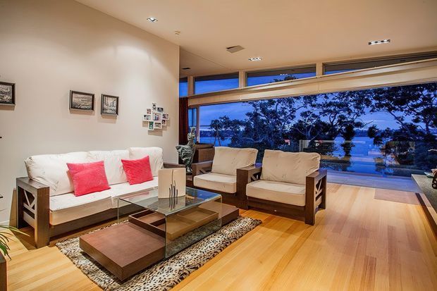A warm, contemporary living room with expansive floor-to-ceiling glazing overlooking trees and water. The room features a white sofa with vivid pink cushions, two upholstered armchairs, a glass coffee table, a low timber ottoman, timber flooring, recessed ceiling lights, and a large patterned rug.