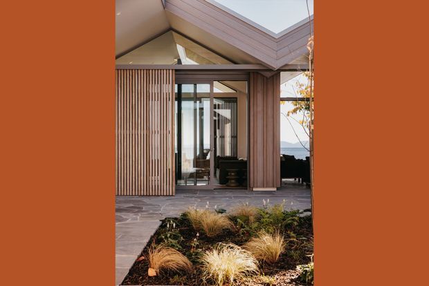 Exterior view of a contemporary timber-clad house with a dramatic pitched roof and large roof skylight or clerestory opening. Vertical timber battens screen the entry and glazing, opening onto a paved terrace with ornamental grasses and a small landscaped garden. Mountainous or coastal scenery is faintly visible beyond the building.