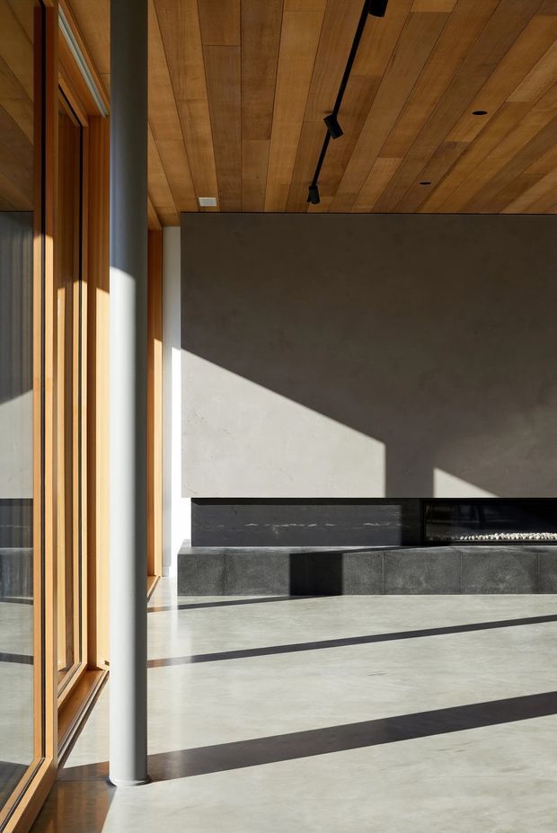 A sunlit contemporary living room centered on a large minimalist fireplace wall. The room features a warm timber-lined ceiling, full-height timber-framed glazing, a white structural column, dark stone or tiled fireplace surround and hearth, polished concrete flooring, and black track lighting. Strong diagonal shadows create a dramatic architectural composition.