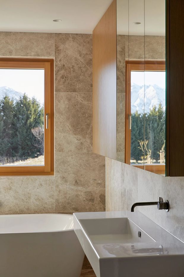 A refined contemporary bathroom with a freestanding white bathtub, integrated rectangular white basin, wall-mounted dark metal tapware, large-format beige stone-look wall tiles, warm timber-framed windows, timber cabinetry, and a mirrored vanity wall. Natural mountain and tree views are visible through the windows and reflected in the mirror.