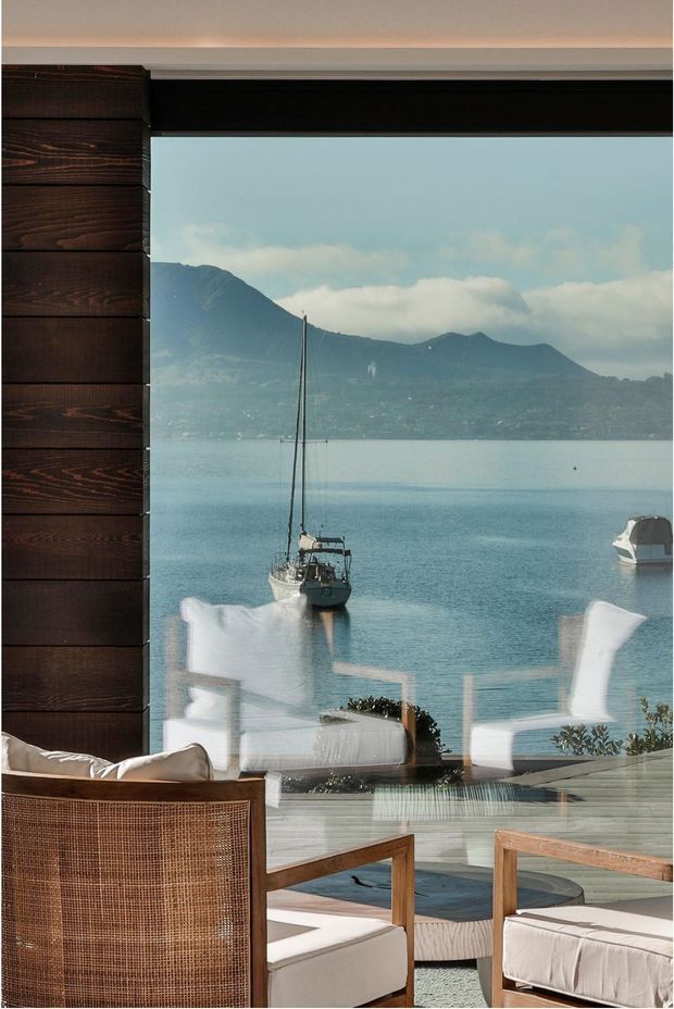 A calm waterfront-facing interior or covered outdoor lounge viewed through large glazing, with dark horizontal timber wall cladding at left and timber-framed, woven-cane lounge chairs with cream cushions in the foreground. The glazing reflects additional white seating. Beyond is a tranquil bay with sailboats and mountainous terrain, creating a strong indoor-outdoor connection.
