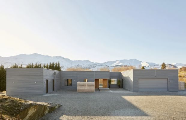 A low, single-storey contemporary residence composed of several interconnected rectangular volumes, viewed from a broad gravel forecourt. The building has horizontal grey cladding, flat roofs, minimal detailing, dark-framed windows and doors, a recessed central entry, and an integrated double garage. Snow-dusted mountain ranges are visible behind the property, with sparse trees and rugged terrain surrounding the site.