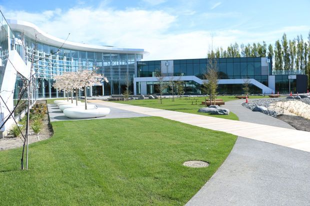 A landscaped modern commercial or institutional campus with a sweeping curved glazed building on the left and a dark contemporary rectangular building in the background. Paved pedestrian paths curve through manicured lawns, with sculptural white seating, young trees, planted areas, and exposed rock landscaping. The scene is bright and open with a clean, contemporary architectural character.