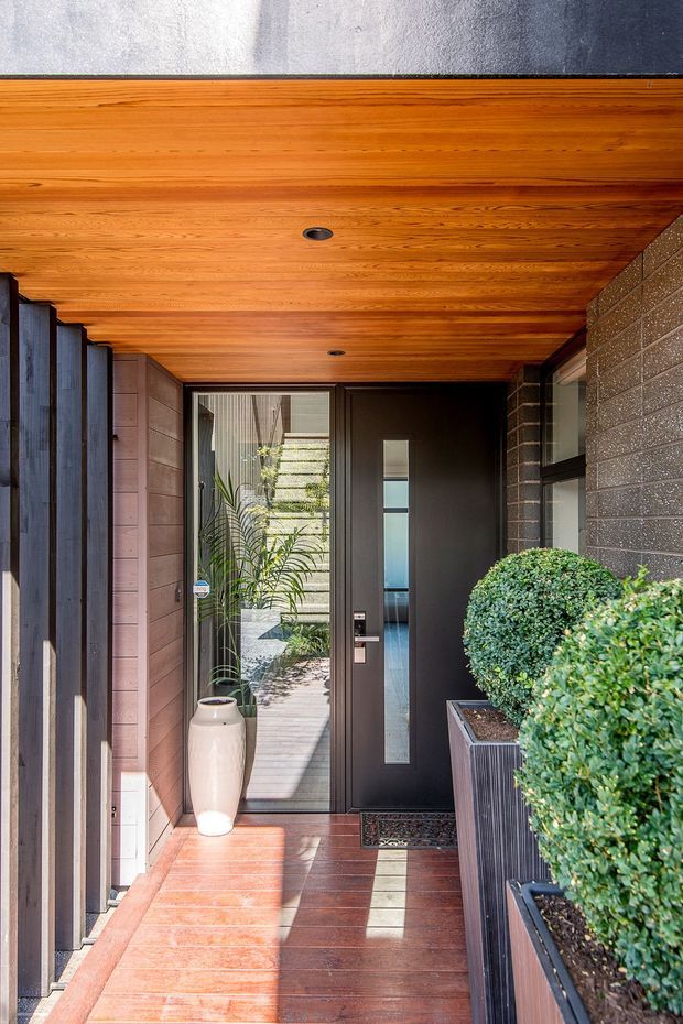 A covered residential entryway with a dark contemporary front door featuring a narrow vertical glazed panel. Warm timber boards line the ceiling and floor, while dark brick and timber-clad walls frame the entrance. Tall glazed openings, a sculptural ceramic planter, and neatly clipped greenery in raised planters create a refined, welcoming approach.