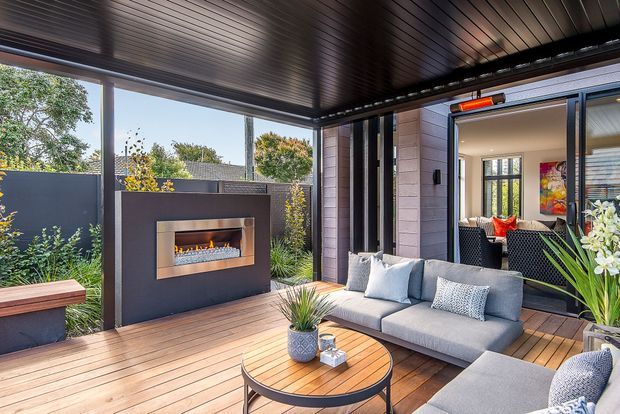 A covered outdoor lounge and entertaining area with timber decking, a built-in linear gas fireplace set into a dark feature wall, a grey outdoor sofa and coffee table, and large sliding glass doors opening into the interior. The space is framed by dark metal elements and a slatted ceiling, with planted garden screening around the perimeter.
