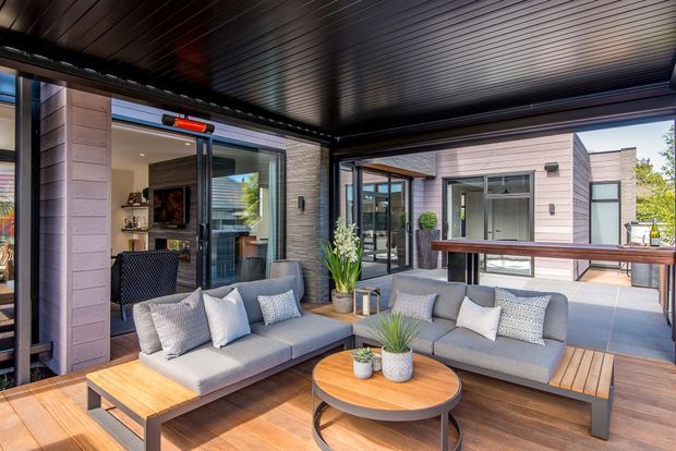 A covered outdoor lounge terrace with two grey cushioned sofas, a timber coffee table, and built-in timber decking. The space is enclosed by dark-framed glazing and a dark slatted ceiling, with a contemporary pale-clad building visible around the courtyard. Potted plants and a raised timber bar counter add function and warmth.