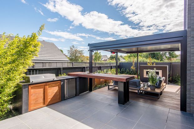A contemporary outdoor entertaining area with a built-in barbecue kitchen, timber-fronted cabinetry, a long dining table, and a covered lounge beneath a modern pergola. Large grey-format paving tiles extend across the terrace, with dark privacy fencing, planted screening, and neighbouring rooftops visible beyond.