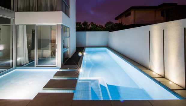 Dusk view of a modern residential pool courtyard, with a long illuminated rectangular swimming pool, tiled steps, dark paving, white rendered boundary walls, and a contemporary glass-fronted multi-storey house. Architectural lighting creates a cool blue and purple atmosphere.