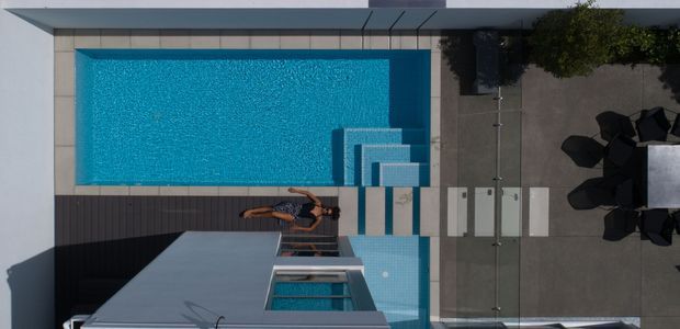 Aerial view of a contemporary residential pool area with a rectangular turquoise swimming pool, integrated shallow step section, dark timber decking, pale tiled paving, and a compact outdoor dining area beside the house.
