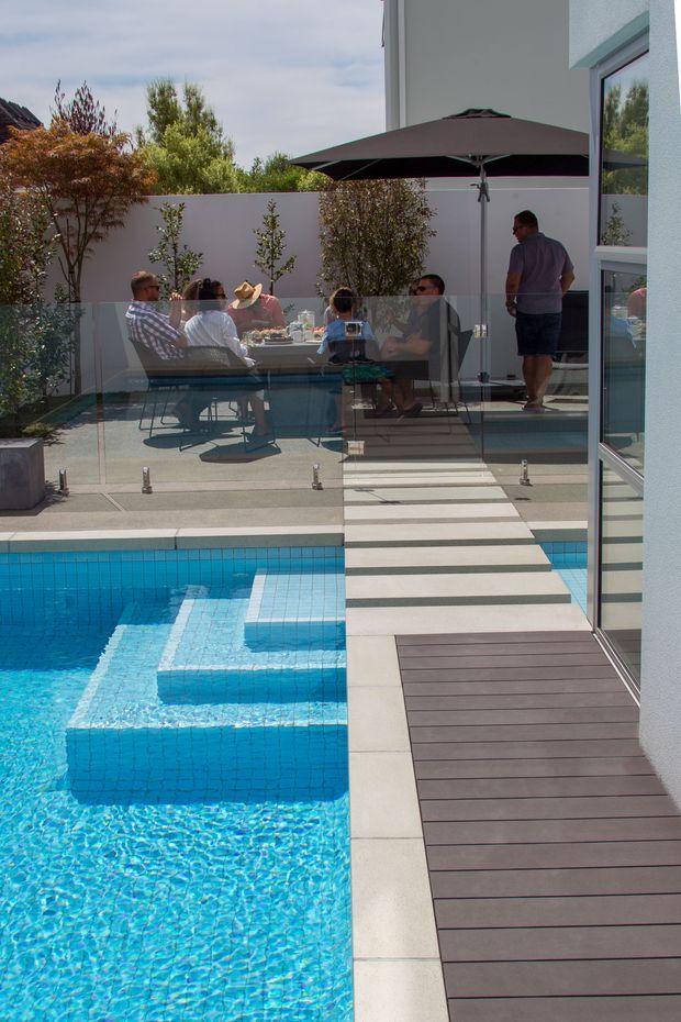 A contemporary outdoor pool area beside a modern white house, featuring vivid blue tiled water, stepped pool sections, dark decking, pale paving, frameless glass balustrades, and a raised entertaining terrace with a dining table and large dark umbrella. Several people are gathered outdoors, with landscaped planting and neighbouring buildings visible in the background.