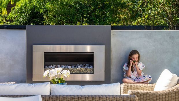 Outdoor lounge area with wicker seating and white cushions arranged against a rendered wall, featuring a built-in stainless-steel fireplace set within a dark feature panel. Lush greenery is visible behind the wall, and a child is seated on the outdoor furniture.