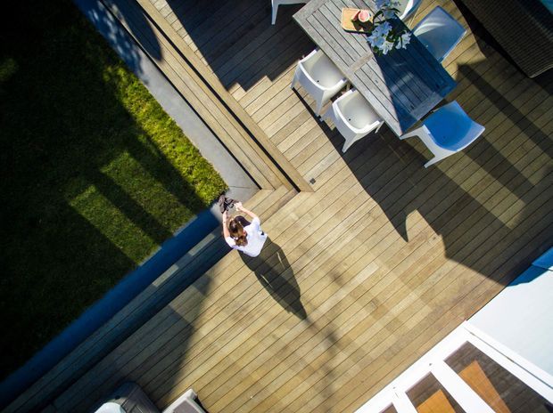 Aerial view of a contemporary timber deck and outdoor dining area beside a narrow linear pool or water feature. The deck includes a dining table with white chairs, a lounge chair, strong geometric shadows, and adjacent lawn and glazed building elements.