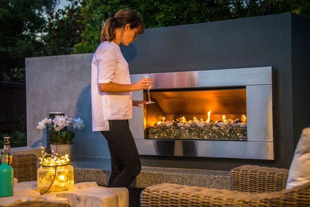 An outdoor entertaining area at dusk featuring a large built-in linear gas fireplace set into a dark grey rendered wall. The fireplace has a stainless-steel surround and a bed of decorative stones with visible flames. Wicker lounge furniture, a stone-topped table, lanterns, drinks, and planting create a warm, relaxed patio setting.