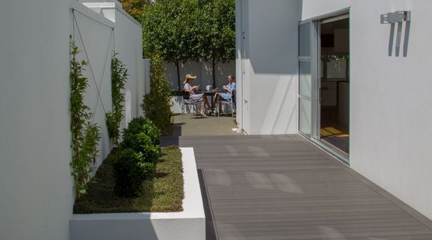 A contemporary outdoor courtyard and deck beside a light-rendered building. The dark timber-look deck leads toward a small seating area with two people, bordered by raised white planter beds, clipped shrubs, and climbing greenery. Large sliding glass doors open into the building, while a minimalist wall-mounted light fixture adds a modern architectural detail.