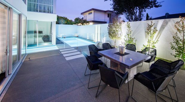 Contemporary outdoor entertaining terrace with a dining table and woven chairs beside a compact swimming pool. The space is enclosed by white rendered walls and surrounded by large glazed openings, with integrated lighting creating a refined evening ambience.
