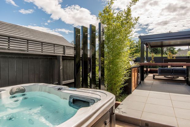 A landscaped residential outdoor entertaining area featuring a bubbling spa pool in the foreground, large-format tiled paving, a covered timber outdoor lounge and dining structure, dark vertical screening panels, and a slatted dark-clad structure. The space has a contemporary, privacy-focused design with greenery and a bright open sky.