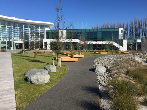 Exterior view of a contemporary institutional or commercial building set within a landscaped public courtyard. The building combines extensive glass curtain walls, dark green-blue cladding, white horizontal elements, and a curved glazed entrance wing. Paved pedestrian paths, timber benches, young trees, lawn, large rocks, and a dry creek-style stone landscape are visible in the foreground.