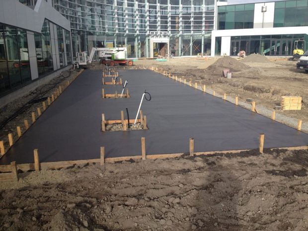 A construction-site view of a newly poured dark concrete slab or paved courtyard between modern commercial buildings. Timber formwork, exposed soil, service conduits, construction equipment and unfinished landscaping are visible, with extensive glass curtain walls and contemporary facades in the background.