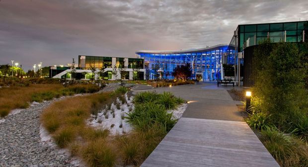 Dusk exterior view of a contemporary institutional or commercial complex, featuring extensive blue-lit glass façades, dark metal framing, landscaped native-style gardens with grasses and gravel, and a timber pedestrian boardwalk leading toward the entrance.