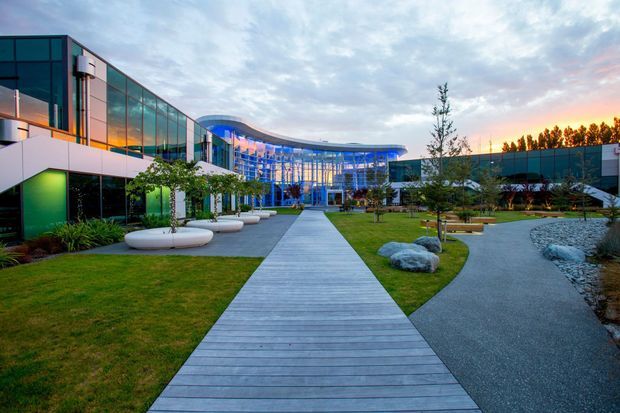 A contemporary commercial campus viewed from a landscaped courtyard, with a broad central timber-look pedestrian boardwalk leading toward a dramatic curved glazed building entrance. The complex features extensive floor-to-ceiling glazing, dark and metallic facade elements, manicured lawns, sculptural rocks, planted areas, curved gravel paths, and integrated outdoor seating beneath an evening sky.