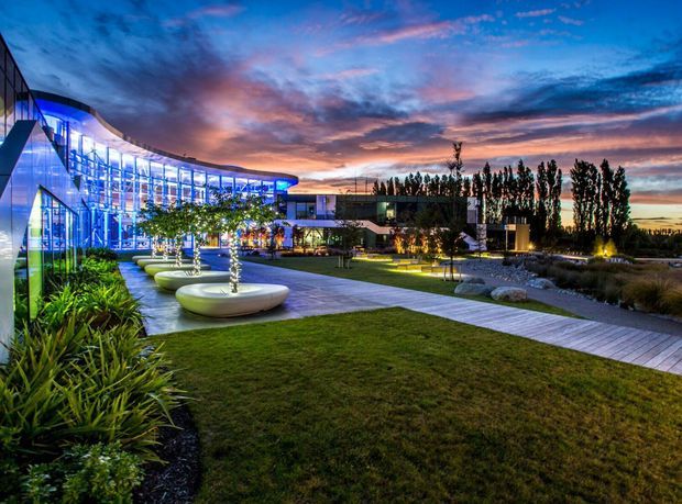 Dusk exterior view of a contemporary commercial or institutional complex with sweeping curved glass façades, dramatic blue architectural lighting, landscaped lawns and garden beds, illuminated ornamental trees, sculptural circular seating or planters, and paved pedestrian paths.