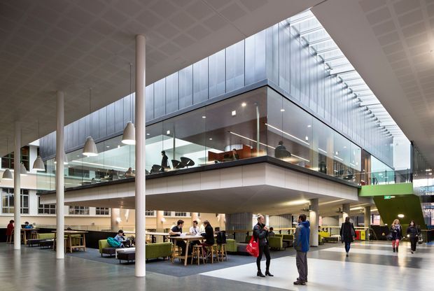 A bright, contemporary multi-level commercial interior resembling an atrium or campus common area. The expansive space features a large glazed mezzanine or upper-level enclosure, exposed white structural columns, a gridded ceiling, extensive clerestory glazing, suspended pendant lights, café-style tables and seating, and people using the communal workspace and dining areas.