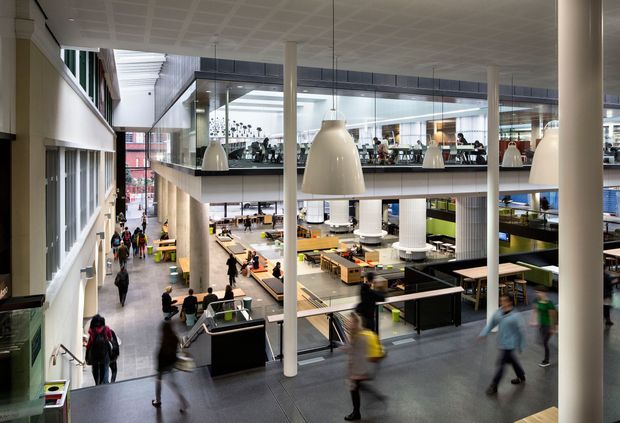 A large, multi-level commercial interior resembling a contemporary food hall, university dining area, or workplace commons. The open atrium contains extensive seating, counters, stairways, balconies, glass partitions, round structural columns, and oversized pendant lights. Numerous people move through the space, while daylight enters through tall glazing and a skylit upper level. The architecture is spacious, functional, and modern with neutral grey, white, black, and timber-toned finishes.