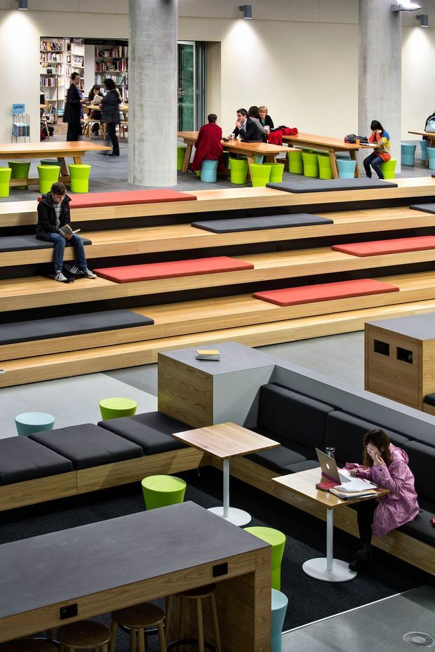 A contemporary multi-level communal interior resembling a library, student lounge, or learning hub. Tiered timber bleacher seating with black and red cushions occupies the centre, while modular timber-and-black upholstered seating, small tables, stools, and colourful lime-green and pale-blue bucket seats create informal work and social areas. Bookshelves, people studying, laptops, concrete columns, and integrated wall lighting are visible in the background.