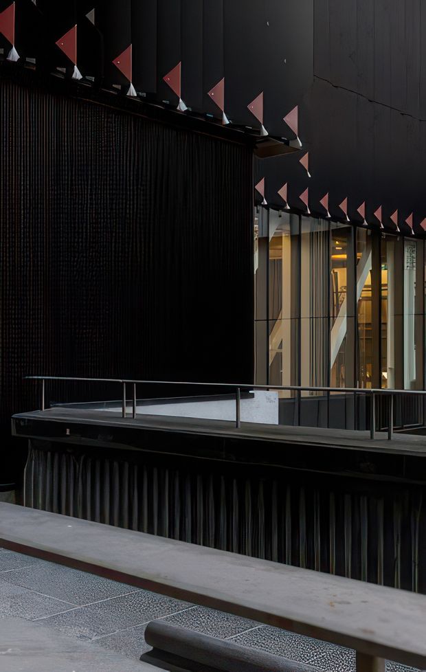 A dark, dramatic contemporary building facade viewed from an elevated walkway or terrace. The architecture features black vertical ribbed cladding, large glazed openings, slim metal railings, and a projecting canopy decorated with repeated pink-red triangular geometric elements. Warm interior lighting is visible through the glazing, while concrete paving and balustrades occupy the foreground.
