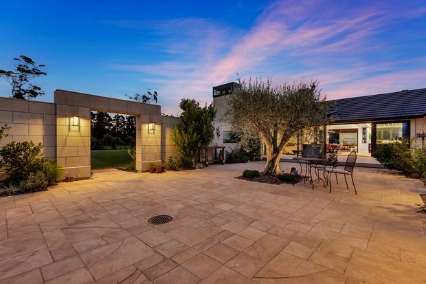 A landscaped residential courtyard or terrace at dusk, featuring a broad paved entertaining area, a mature olive tree, outdoor dining furniture, planted borders, and a contemporary low-rise building with warm interior lighting. A rendered masonry gateway with integrated wall lights opens toward a lawned garden beyond.