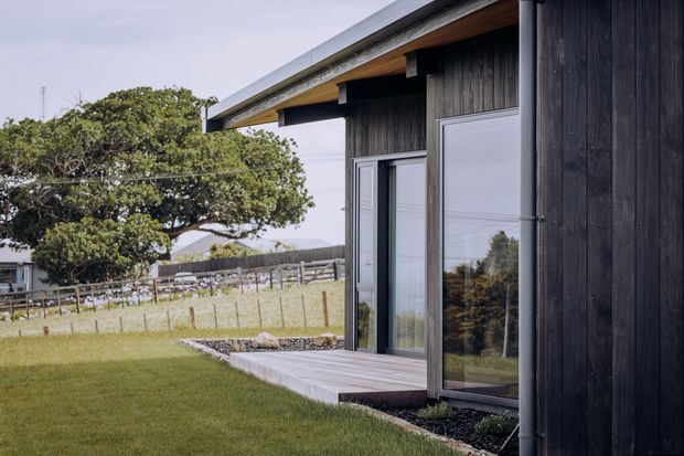 Exterior detail of a contemporary single-storey building with dark vertical timber cladding, extensive glazing, exposed timber eaves, and a low-pitched roof. A small timber deck and concrete threshold open onto a landscaped lawn, with a rural fenced setting and mature tree beyond.