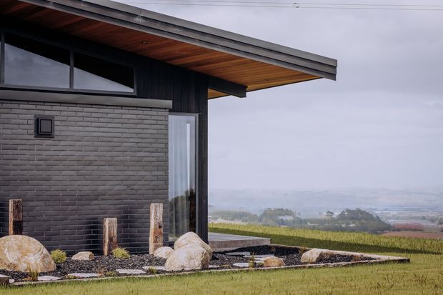 Exterior view of a contemporary single-storey building set in an open rural landscape. The facade combines dark vertical cladding, grey brickwork, expansive dark-framed glazing, and a broad projecting roof with a warm timber soffit. A minimalist landscaped foreground uses gravel, large natural boulders, timber posts, and low planting, with expansive farmland and distant hills beyond.
