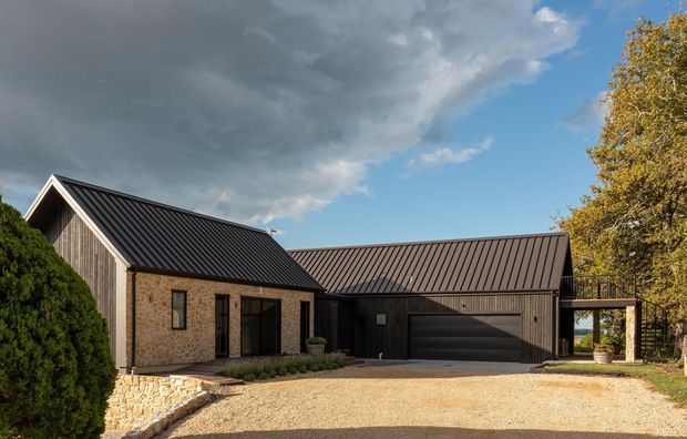Exterior view of a low-profile contemporary rural residence with a gabled stone-clad wing connected to a dark metal-clad garage or living wing. The building has dark standing-seam metal pitched roofs, black-framed glazing and garage doors, a gravel driveway, a low stone retaining wall, and mature landscaping.