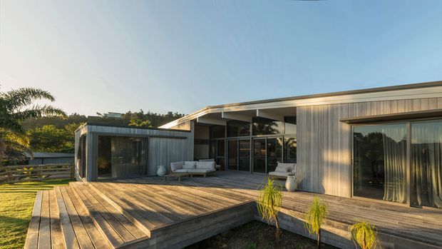 Single-storey contemporary residence viewed from the rear, with a broad timber deck wrapping around the house, extensive dark-framed glazing, and vertically clad timber or wood-look exterior walls. The low, gently sloping roof and open indoor-outdoor living arrangement face a landscaped garden in warm late-day light.