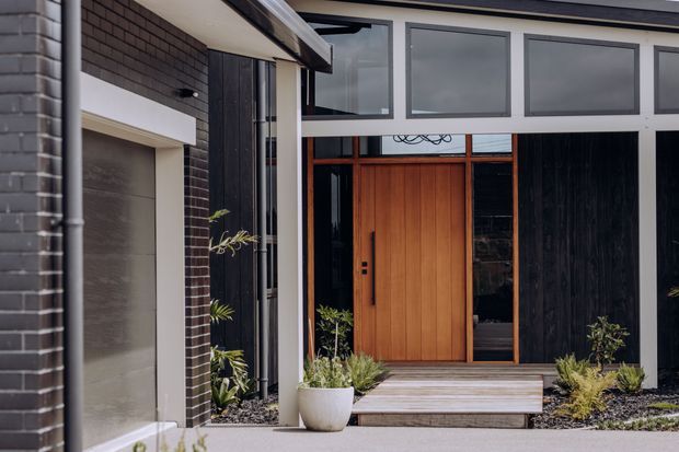 A contemporary residential front entry viewed from outside. The facade combines dark brick masonry, black vertical cladding, large framed glazing, and a warm timber front door. A shallow covered canopy leads to broad timber steps, with minimalist landscaped gravel beds and potted plants framing the entrance.