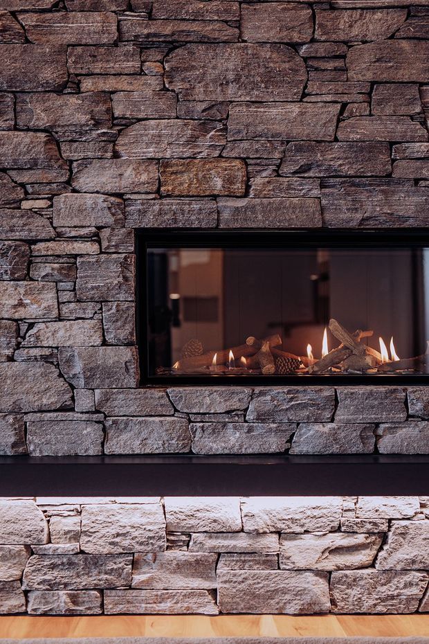 Close-up interior view of a contemporary stacked-stone feature wall with a long, black-framed linear fireplace inset into the masonry. The fireplace contains decorative logs and visible flames, with a darker horizontal ledge below and timber flooring at the base. The image focuses on textured natural stonework and the warm glow of the fire.