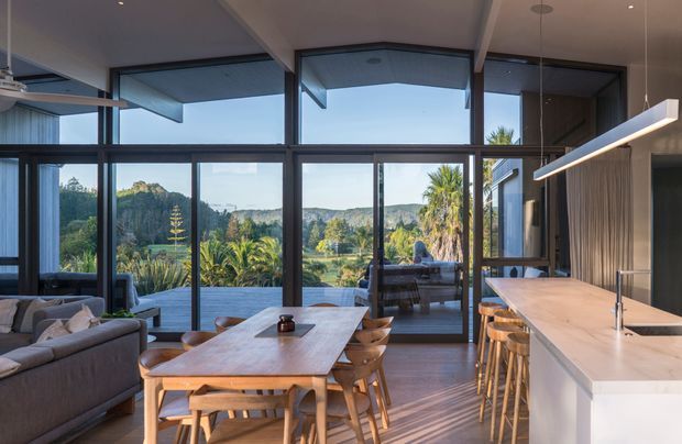 Open-plan contemporary kitchen, dining and living area with floor-to-ceiling glazing overlooking a lush, hilly landscape. A large light timber dining table with matching chairs occupies the centre, while a pale kitchen island with timber stools and an integrated linear pendant light runs along the right side. Dark window and door frames, timber flooring, exposed ceiling beams and an outdoor deck are visible.