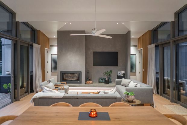 A contemporary open-plan living and dining area viewed from the dining table. The room features a large timber dining table, upholstered sectional sofa, built-in fireplace and television set into a dark textured feature wall, timber wall panelling, extensive dark-framed glazing, recessed ceiling lights, and a central ceiling fan. The space is bright, refined, and connected to outdoor areas through large glazed doors.