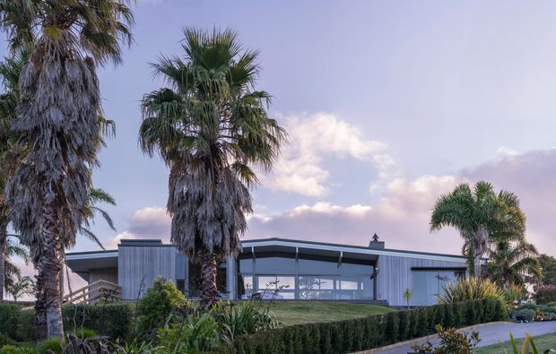Exterior view of a low-slung contemporary residence with a flat roof, extensive floor-to-ceiling glazing, and light vertical metal or timber-like cladding. The building is framed by tall palm trees, clipped hedges, lush landscaping, and a paved approach in soft evening light.