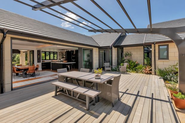 A bright, contemporary courtyard entertaining area with a large timber deck, outdoor dining table and bench seating, and an open pergola structure overhead. The courtyard is enclosed by single-storey wings with grey tiled pitched roofs, light masonry walls, extensive sliding glass doors, and a visible indoor kitchen and dining area. Lush built-in planting adds a natural accent around the perimeter.