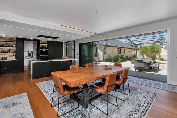 Contemporary open-plan kitchen and dining space with dark cabinetry, a central island, warm timber flooring, and a large solid timber dining table surrounded by leather dining chairs. Full-height glazed doors open to a paved courtyard with a pergola, bringing in abundant daylight.